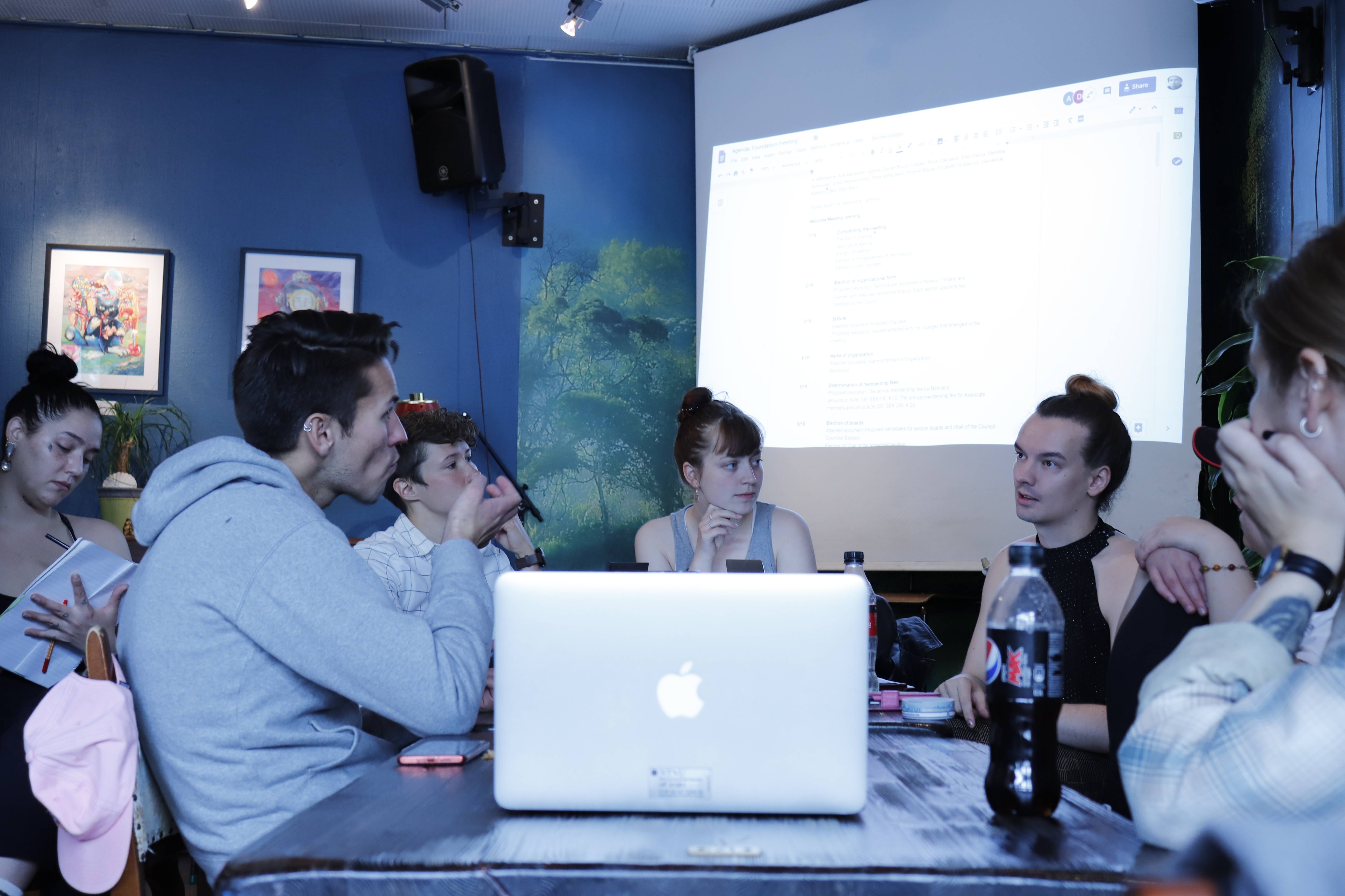 A group of people having a meeting, sitting around the same table, with a projector and a person taking notes in the background.